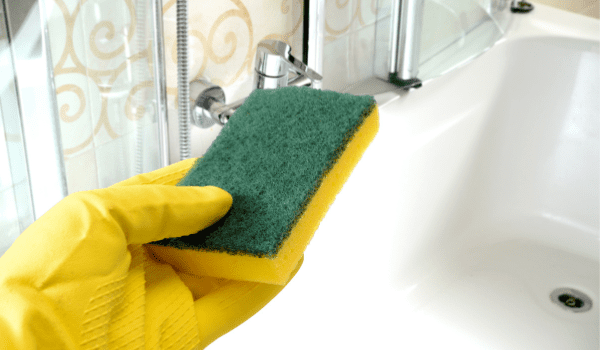 Image of a hand wearing a yellow glove and holding a sponge, with a bath in the background.