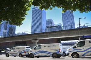 Metro Plumb vans driving on the road, with a large city skyline in the background, representing mobile plumbing services.